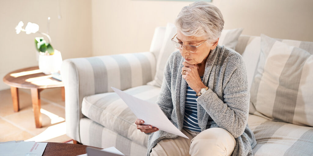 Une femme âgée aux cheveux blancs, assise sur un canapé, consulte attentivement un document relatif à sa pension de réversion après le décès de son conjoint.