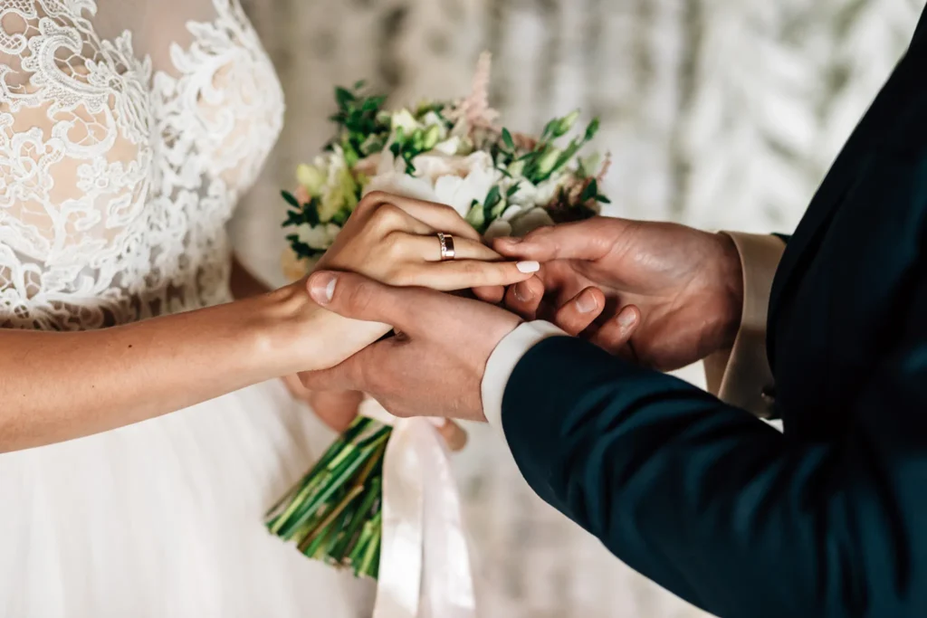 Photo gros plan des mains d'un couple fraîchement marié échangeant leurs alliances lors de la cérémonie. La main de la mariée, portant une bague en or, tient un bouquet de mariée, tandis que la main du marié la soutient doucement. Cette image symbolise l'union et l'engagement, faisant écho à l'importance de la protection mutuelle et des décisions éclairées comme le contrat de mariage.